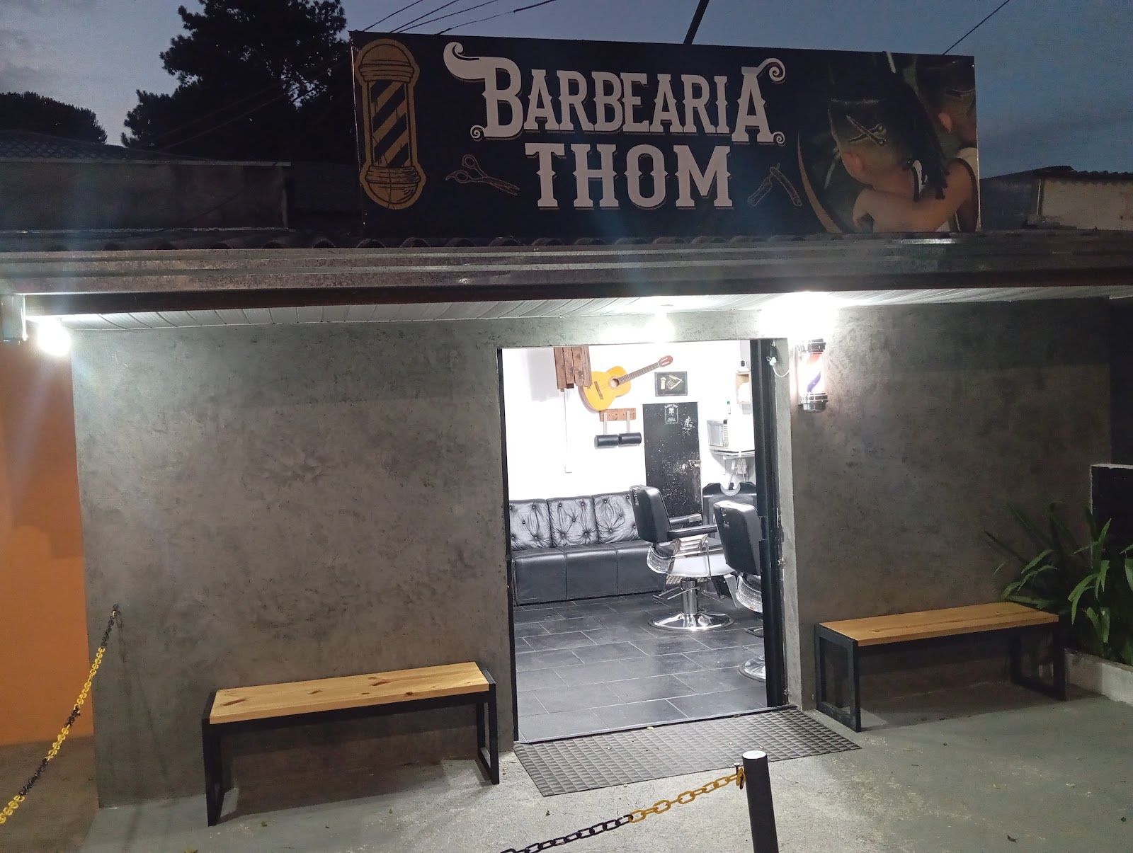 Fachada iluminada da Barbearia Thom à noite, com letreiro e poste de barbeiro na entrada